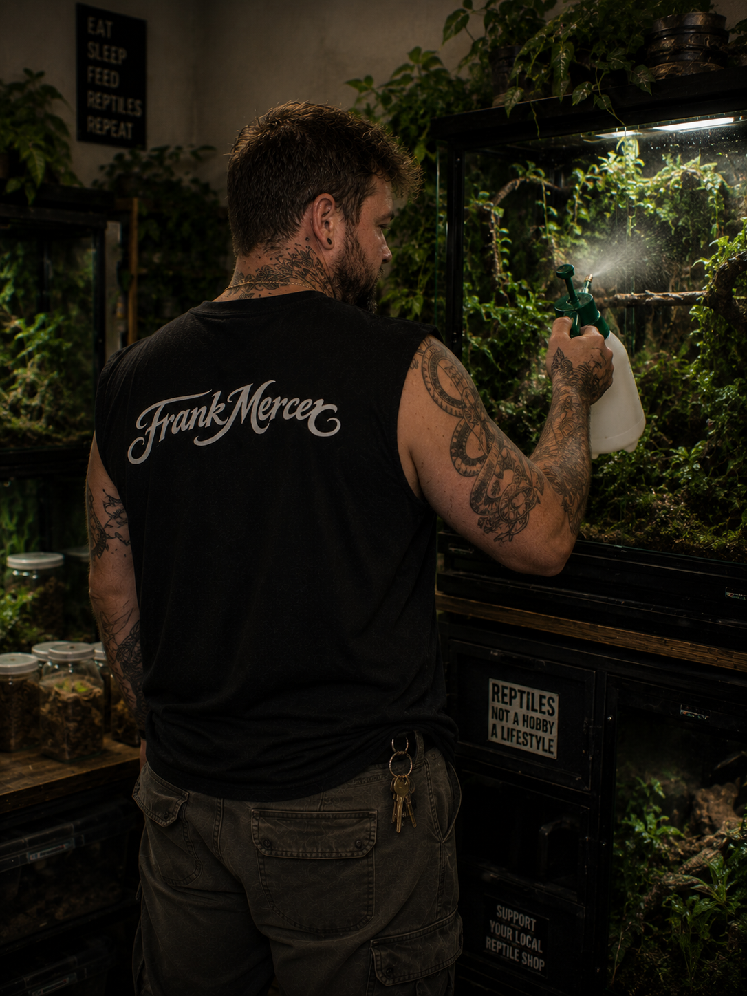 Man in a black 'Frank Mercer' shirt holding a bottle in a dimly lit room with plants.